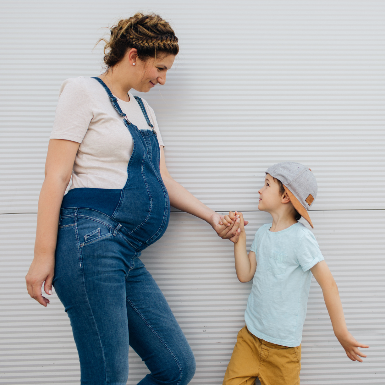 Cute pregnant mom in overalls holding hands with her elementary age son against a white wall.
