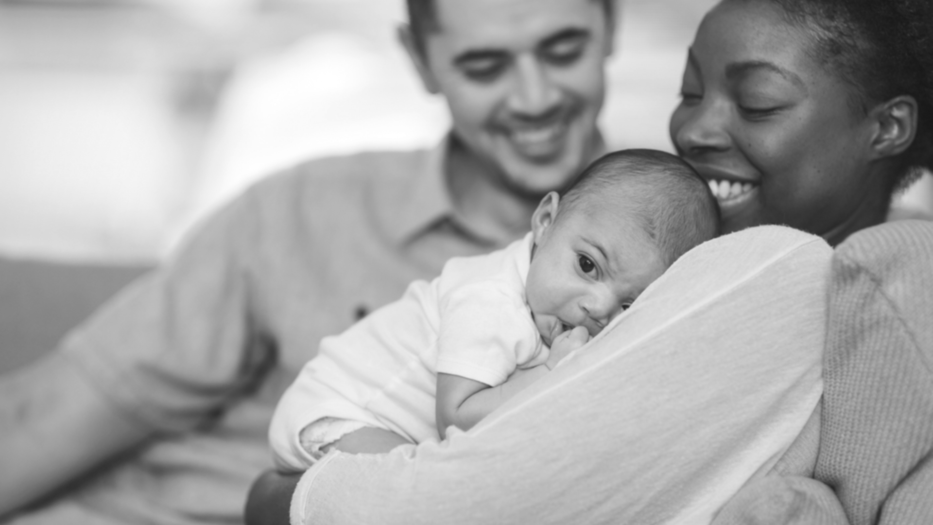 Black and white image of new family. Happy dad looks on while happy mom holds infant baby. Birth Order Bundles of Joy.
